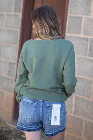 A woman viewed from behind wearing an olive green long-sleeve top and blue denim shorts with a visible white Levi's tag on the back pocket, standing against a gray brick wall.