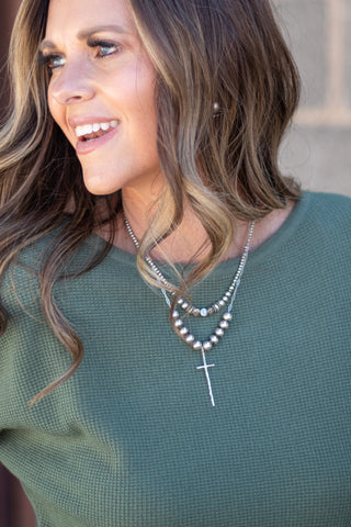 A close-up lifestyle shot of a woman wearing a silver multi-strand necklace with a cross pendant over a textured olive green top.