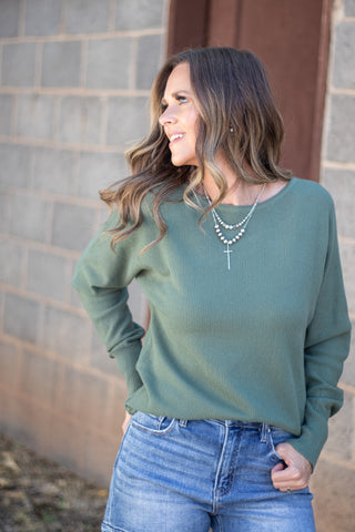 A woman wearing a sage green long-sleeve knit top and blue jeans, standing outdoors against a gray brick wall.