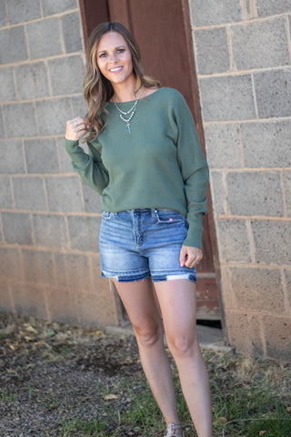 A woman wearing a sage green long-sleeve knit top and denim cutoff shorts, standing outdoors in front of a gray brick wall.