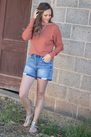 A woman smiling and posing outdoors, wearing a long-sleeve crewneck sweater in a muted terracotta or burnt orange color, paired with blue denim shorts.