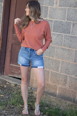 A woman stands outdoors against a gray cinderblock wall and brown wooden door, wearing a rust-orange long-sleeve crewneck top, light-wash denim cutoff shorts, and clear high-heel sandals.
