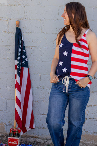 Woman wearing a tank top with an American flag design, standing next to a brick wall and American flag.