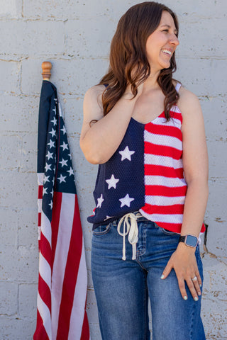 Woman wearing an American flag tank top standing next to an American flag against a white wall.