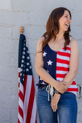 Woman wearing a tank top with American flag design standing next to an American flag against a light gray wall.