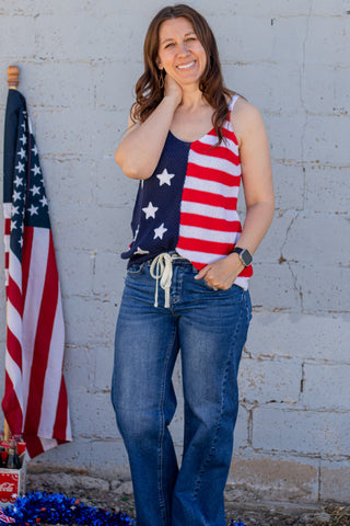 Woman wearing a tank top with an American flag design, standing next to a brick wall and American flag.