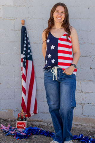 Woman wearing a tank top with an American flag design, standing next to a brick wall and American flag.